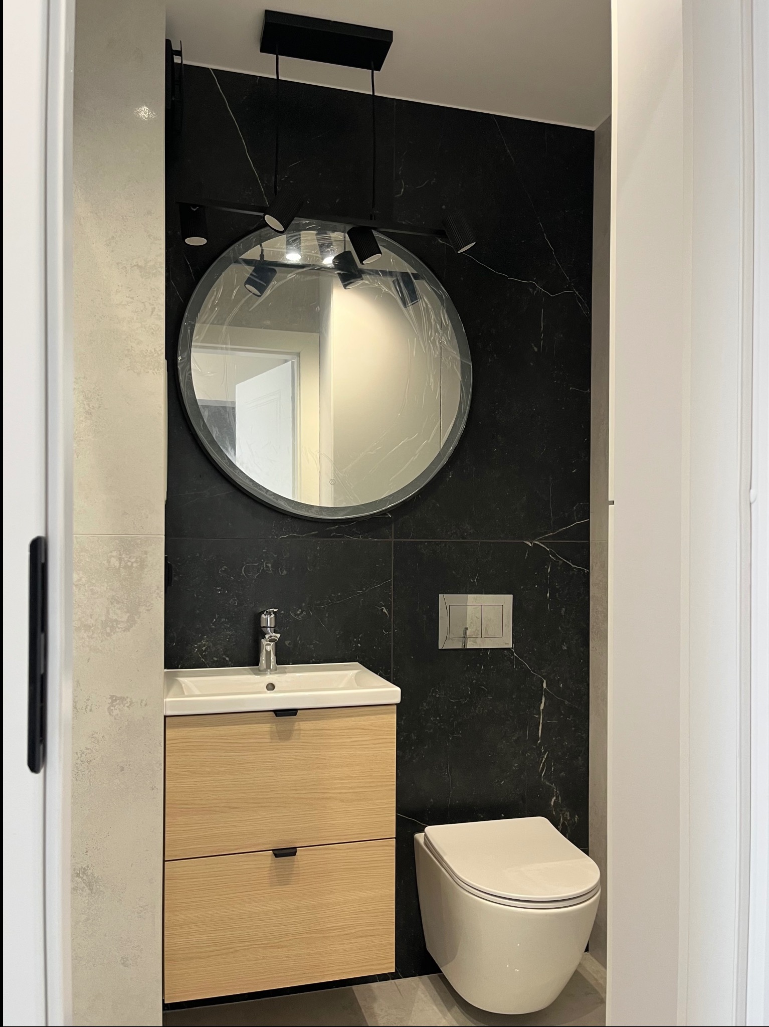 Nowoczesna łazienka z czarną ścianą imitującą marmur, okrągłym lustrem z folią ochronną, drewnianą szafką pod umywalką i podwieszaną toaletą. Minimalistyczny design.