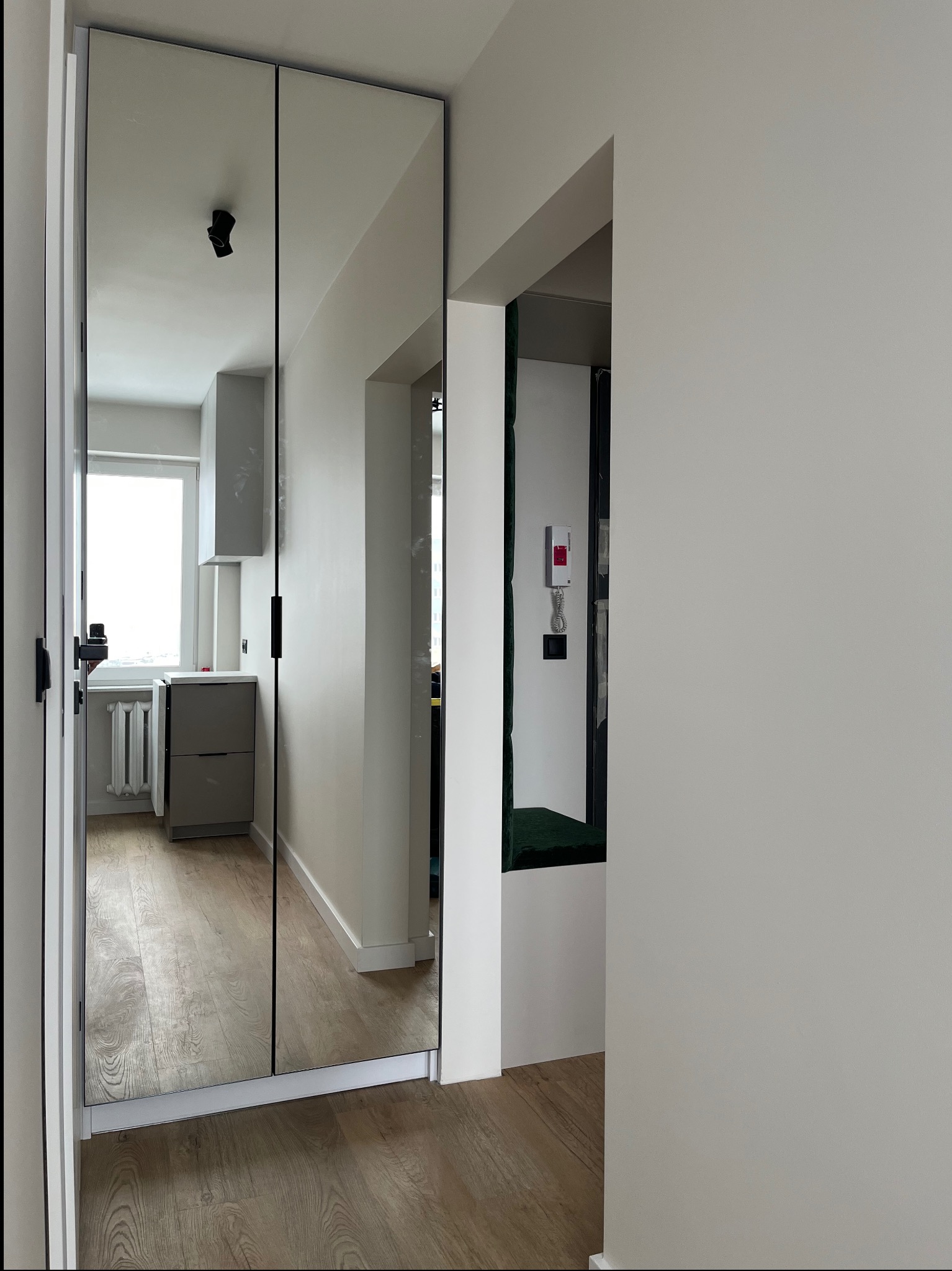 Nowoczesna szafa wnękowa z lustrzanymi drzwiami, minimalistyczne uchwyty, widok na jasną kuchnię w tle, drewniana podłoga, białe ściany.