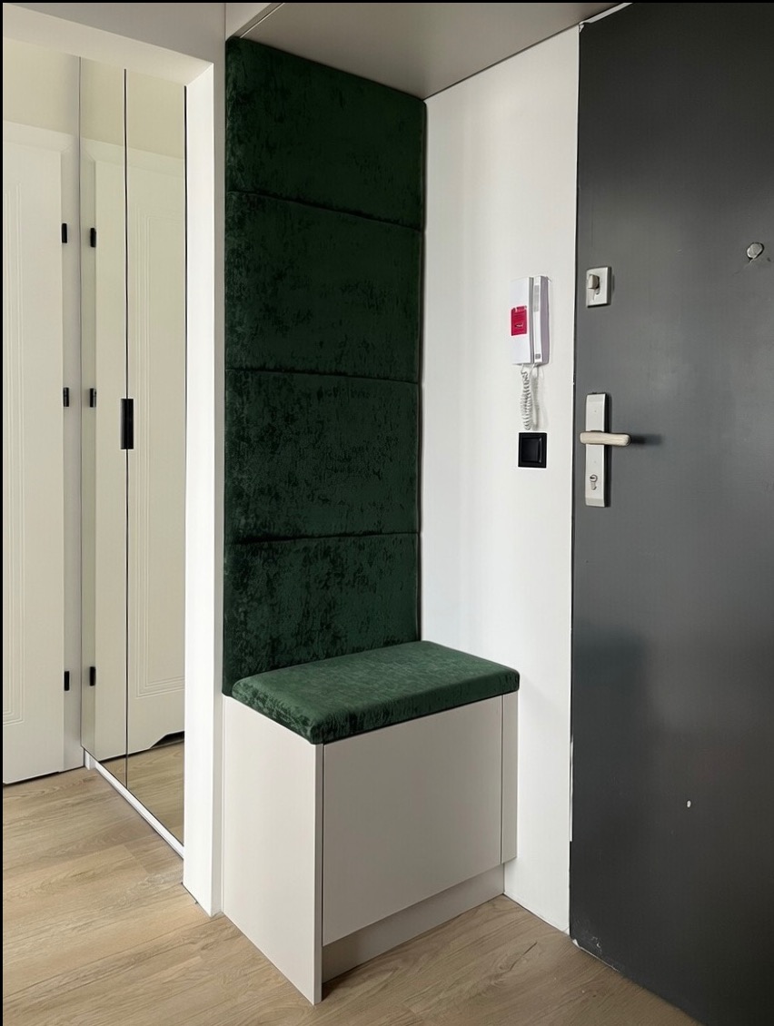 Nowoczesna zabudowa przedpokoju: biała szafka z siedziskiem obitym butelkową zielenią, panele ścienne w tym samym kolorze. Obok drzwi wejściowe i lustrzana szafa.