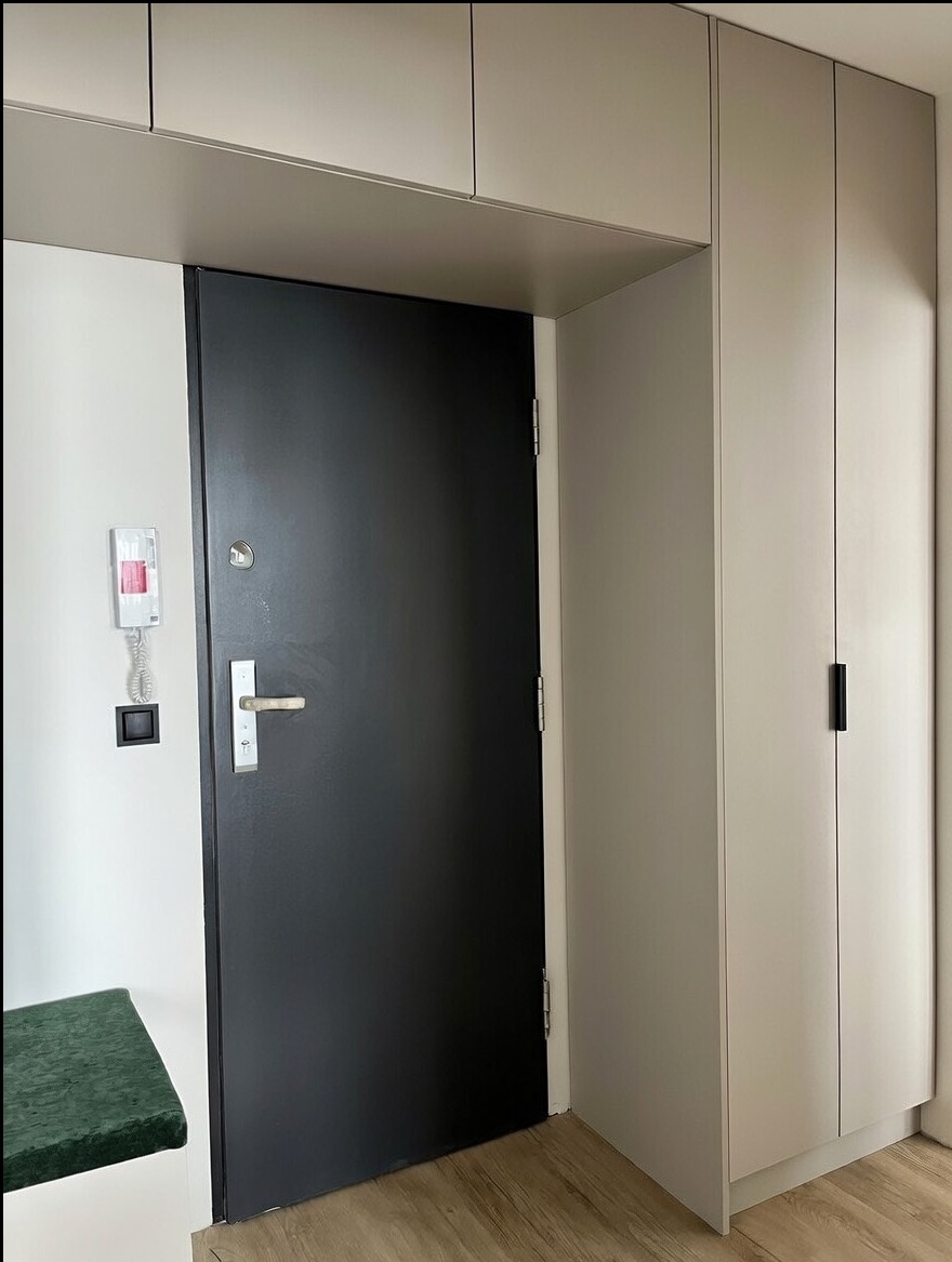 Nowoczesne wykończenie przedpokoju: czarne drzwi wejściowe, zabudowa szafy w odcieniach szarości, zielone siedzisko. Minimalistyczny design, jasne panele podłogowe.