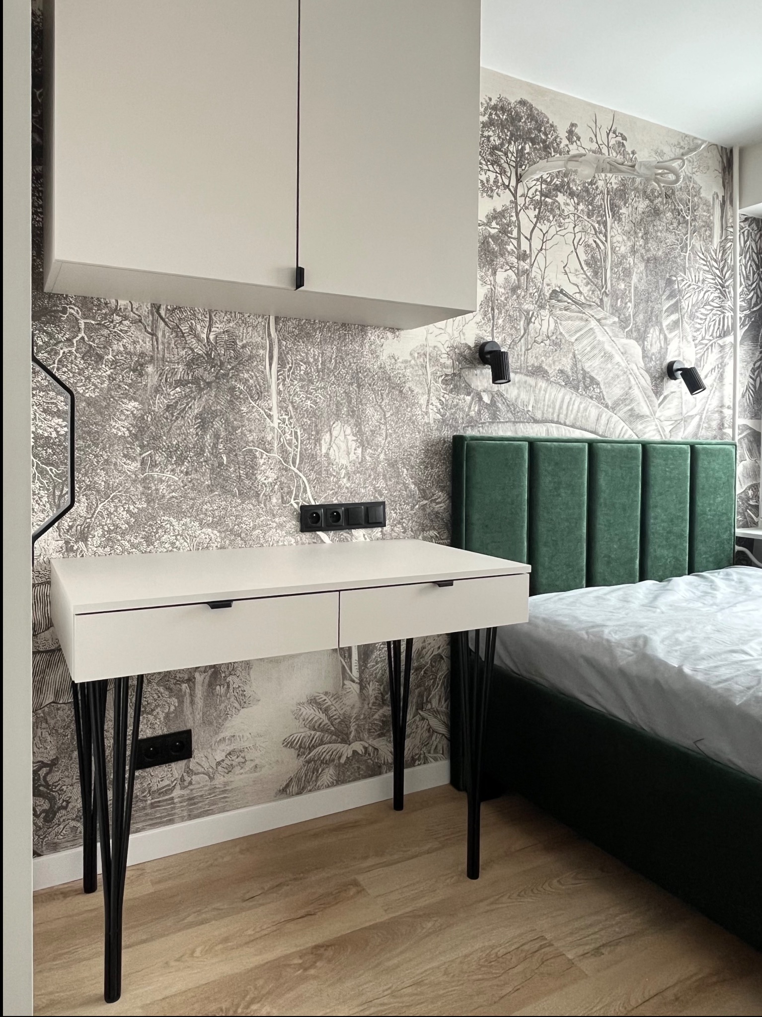 Białe biurko z szufladami na czarnych, metalowych nogach w sypialni z tapetą w motywy roślinne i zielonym zagłówkiem łóżka. Nowoczesny design.