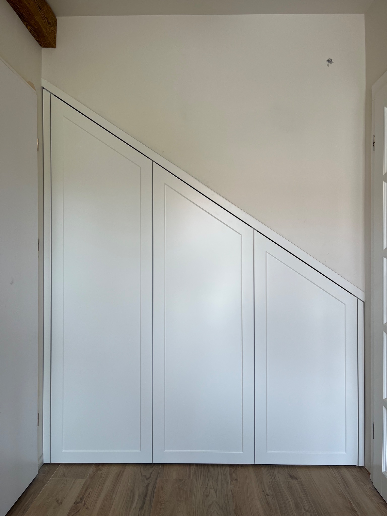 Biała szafa wnękowa pod skosem na poddaszu, z drewnianą belką stropową w rogu. Minimalistyczny design, panele z delikatnym frezem.