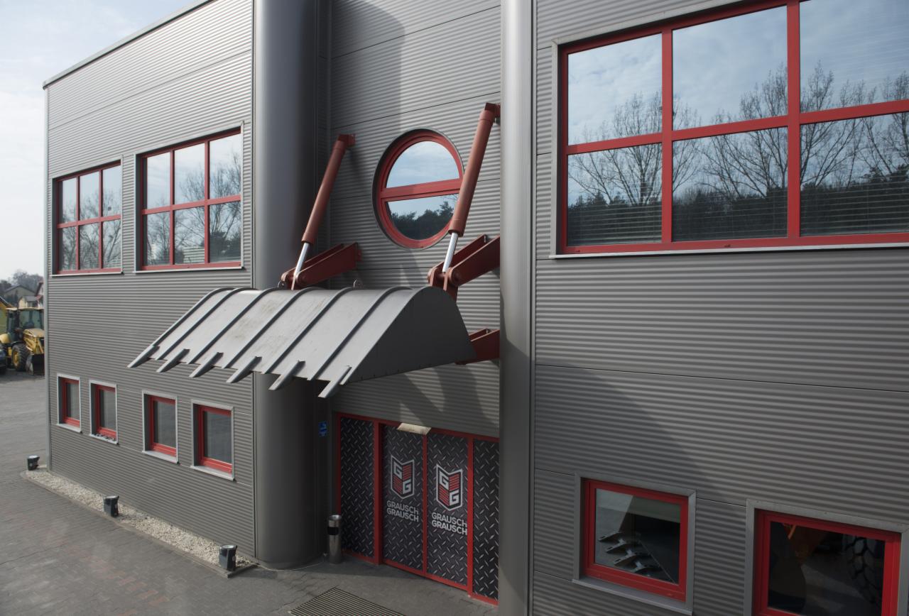 Fasada budynku firmy z elementami dekoracyjnymi nawiązującymi do maszyn budowlanych: stylizowana łyżka na wysięgnikach nad wejściem, okrągłe okno, szare blaszane panele ścienne i czerwone ramy...