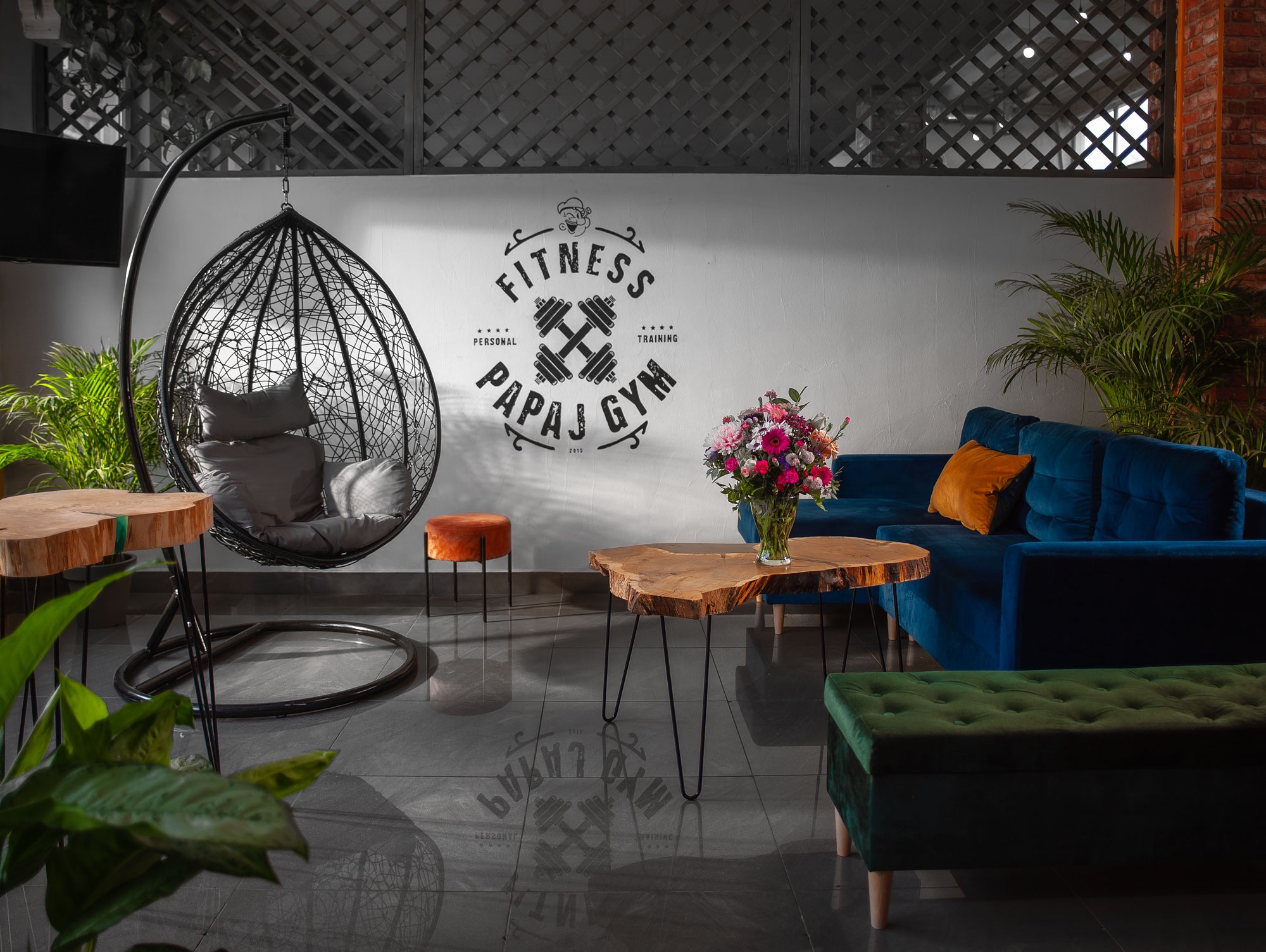 Wnętrze siłowni Papaj Gym z huśtawką w kształcie kokonu, drewnianym stolikiem z bukietem kwiatów, niebieską sofą i zielonym pufem, logo na ścianie.