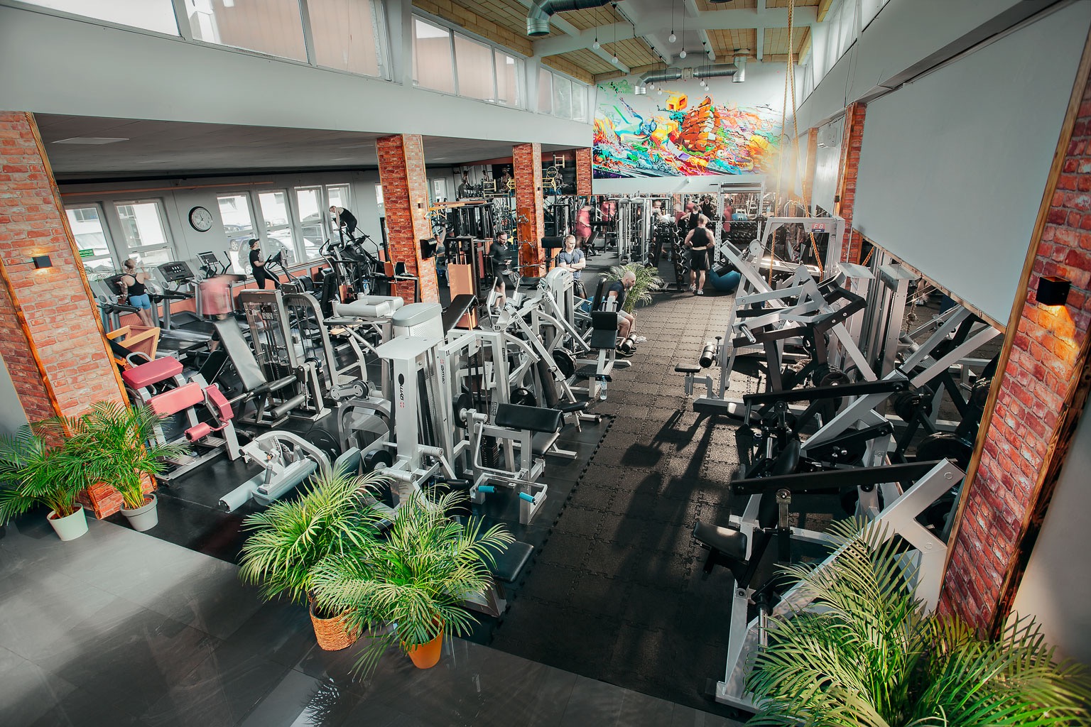 Widok z góry na salę ćwiczeń w Warszawie z różnorodnym sprzętem do ćwiczeń siłowych i cardio, z osobami trenującymi na bieżniach i maszynach, dekorowana cegłą i kolorowym muralem.