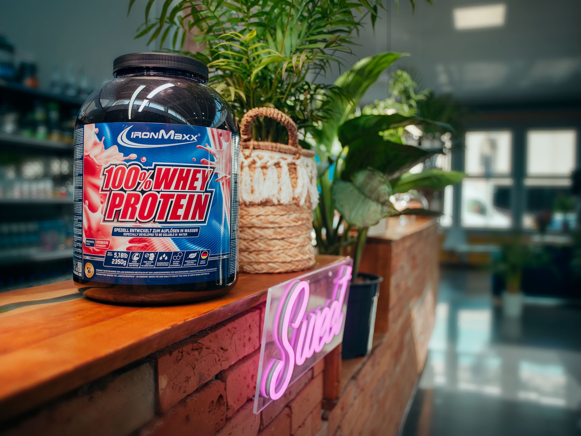 Pojemnik Ironmaxx 100% Whey Protein o smaku truskawkowym, umieszczony na blacie z ceglanym wykończeniem, obok neonowego napisu 'Sweet' i roślin doniczkowych.