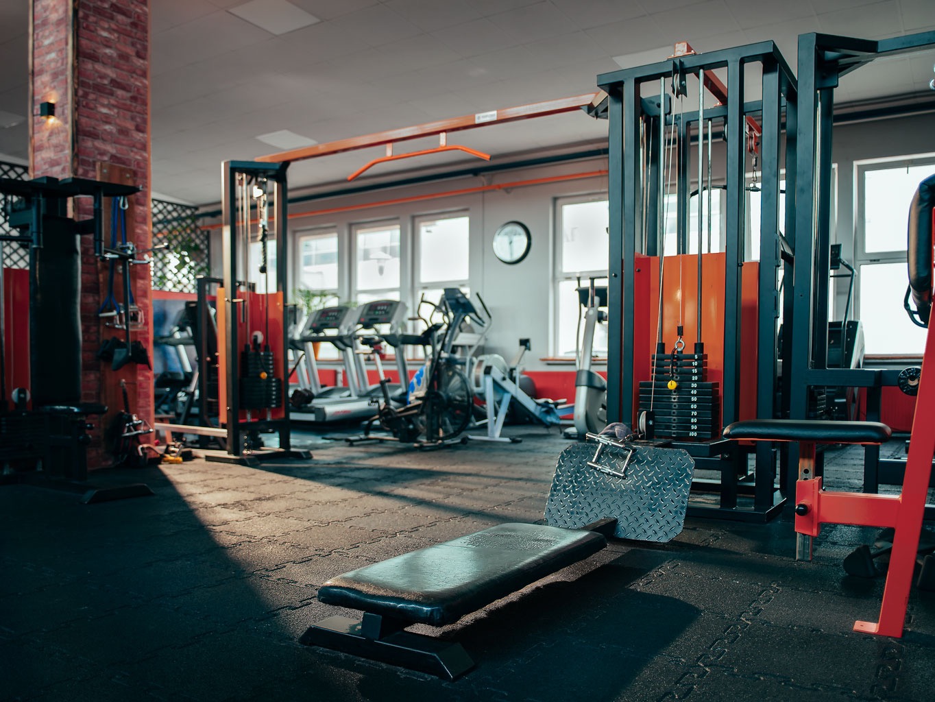 Wnętrze siłowni z maszynami do ćwiczeń, bieżniami i rowerami treningowymi w tle, widok z niskiej perspektywy na matę podłogową i część maszyny do ćwiczeń.