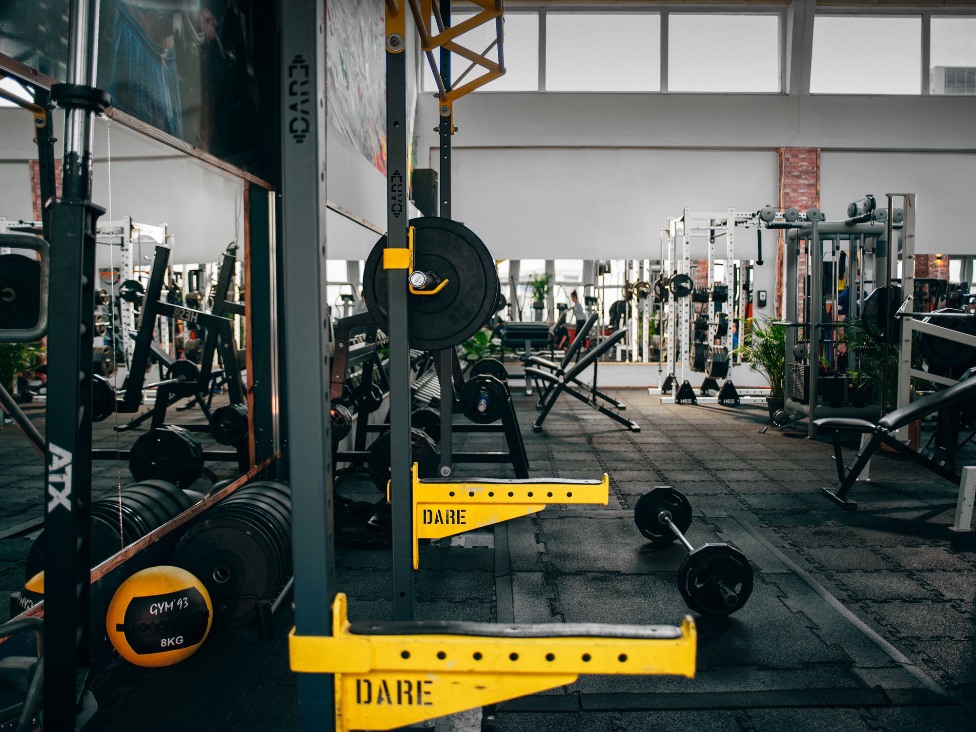 Wnętrze siłowni z widocznym sprzętem do ćwiczeń: stojaki z obciążeniami, sztangi, 8kg piłka lekarska, lustro oraz żółte uchwyty z napisem DARE.