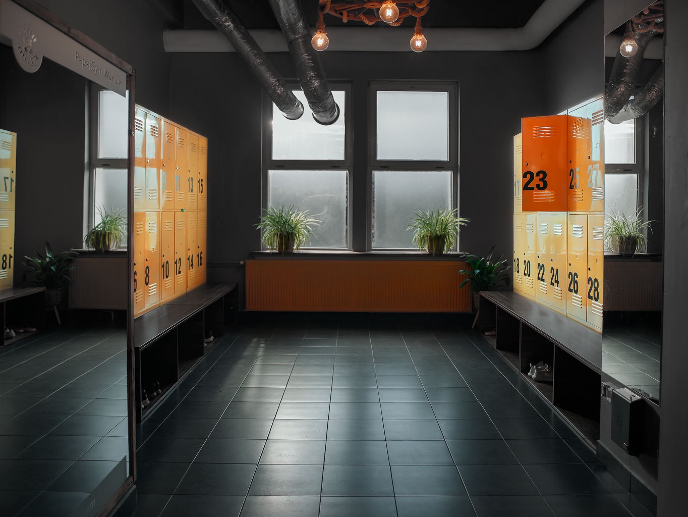 Szaro-pomarańczowa szatnia w siłowni Papa Gym Warsaw z rzędami szafek z numerami, ławkami, roślinami doniczkowymi i oknami w tle, oświetlona lampami z żarówkami Edisona.