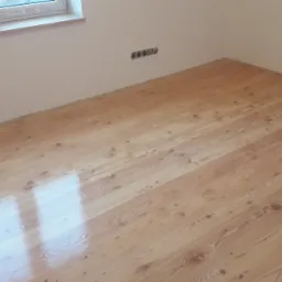 Świeżo polakierowany parkiet z jasnego drewna sosnowego, widoczne ślady pędzla i refleksy światła, gładkie ściany w tle, gniazdka elektryczne na ścianie.