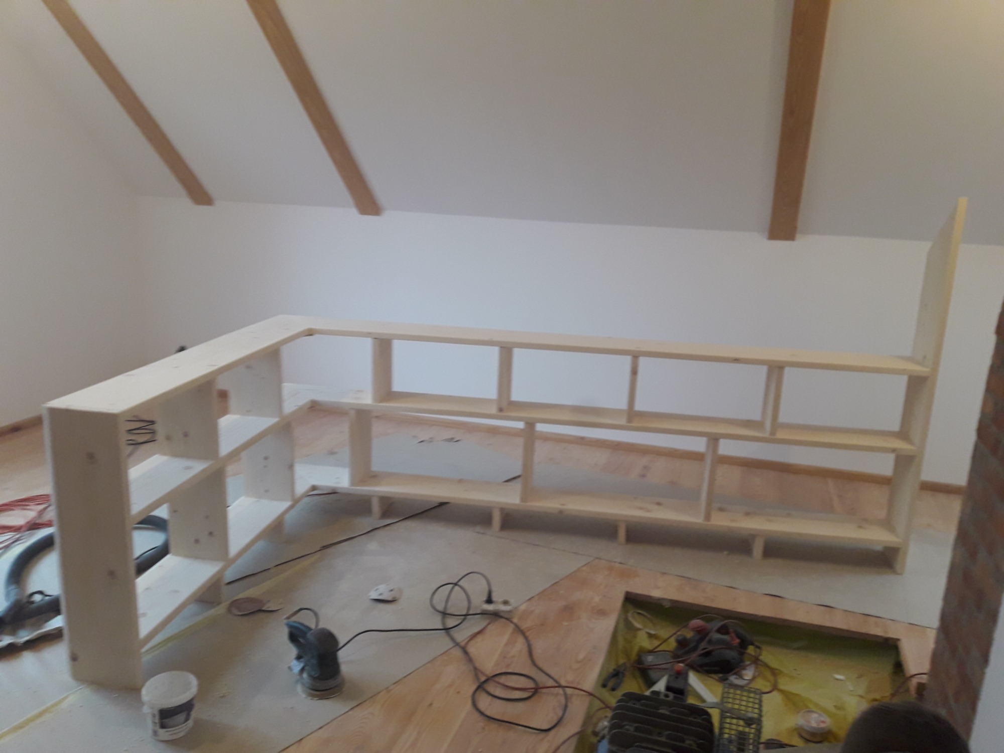 Montaż drewnianego regału na poddaszu ze skośnym sufitem, widoczne narzędzia i nieukończona podłoga.