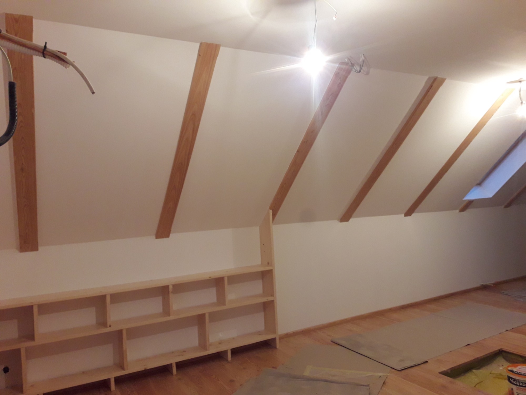 Poddasze w trakcie wykończenia: drewniane belki konstrukcyjne, jasne ściany, drewniana zabudowa regału, zabezpieczona podłoga i widoczne przewody instalacji elektrycznej.
