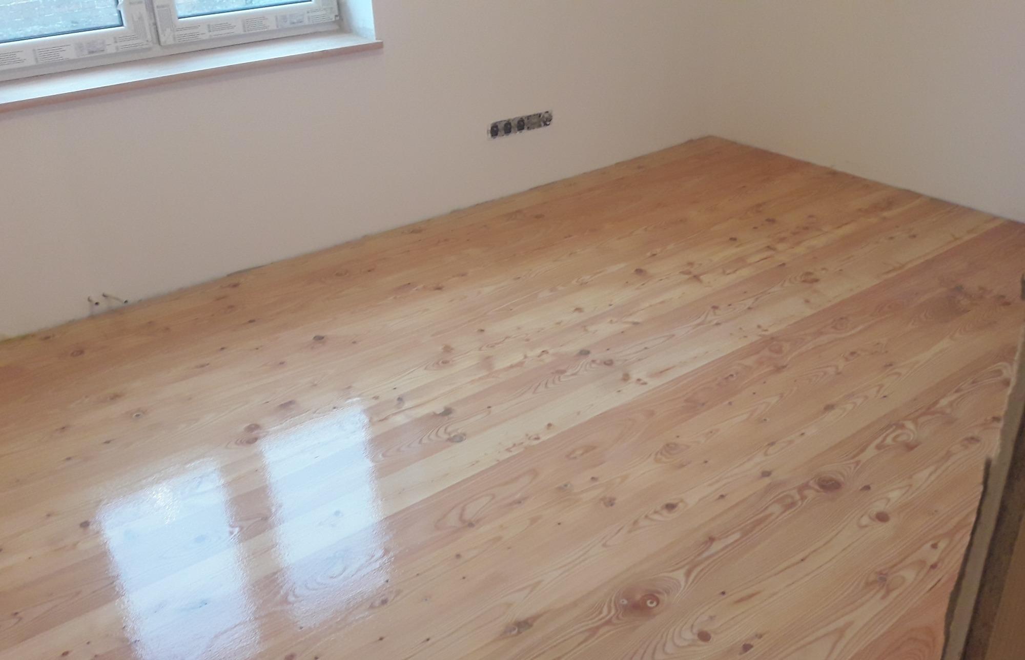 Świeżo polakierowany parkiet z jasnego drewna sosnowego, widoczne ślady pędzla i refleksy światła, gładkie ściany w tle, gniazdka elektryczne na ścianie.