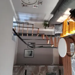 Wnętrze mieszkania z drewnianymi schodami spiralnymi z czarnymi poręczami, szarym dywanem w marokański wzór, białym stolikiem kawowym i kanapą z żółtą narzutą oraz śpiącym psem.