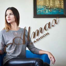 Kobieta siedzi na niebieskim krześle, ubrana w szary sweter z ażurowym dekoltem i czarne sk&oacute;rzane spodnie, na ścianie wisi obraz z motywem weneckim, widoczne logo marki 'Almax Fashion'.