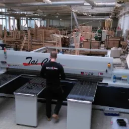 Pracownik obsługuje piłę panelową Tai CNC 105H w warsztacie, w tle składowane drewno i okna z widokiem na zewnątrz.