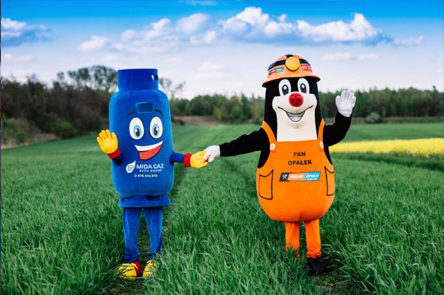 Dwie maskotki reklamowe: niebieska butla z gazem z uśmiechem i kret w stroju górnika, stojące na zielonym polu i trzymające się za ręce.
