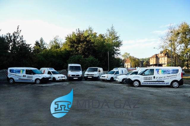 Flota pojazdów dostawczych z logo firmy dostarczającej gaz, zaparkowana na placu z drzewami w tle.