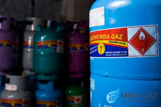 Stos butli z propan-butanem różnych kolorów, z niebieską butlą na pierwszym planie, na której widoczna etykieta z informacjami o produkcie i ostrzeżeniami.