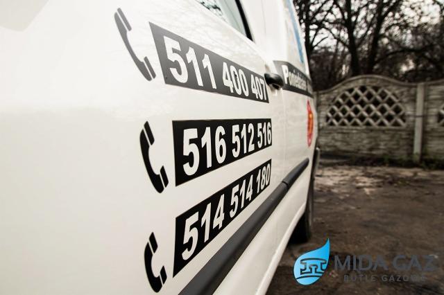 Biały samochód dostawczy z czarnymi naklejkami z numerami telefonów i logo firmy Mida Gaz, zaparkowany na tle betonowego ogrodzenia i drzew bez liści.
