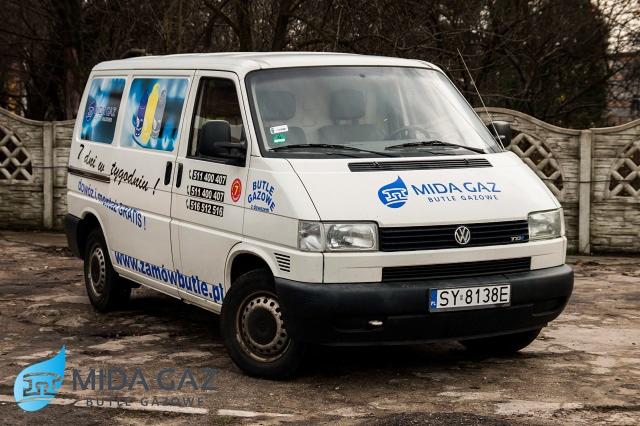 Biały samochód dostawczy z logo firmy Mida Gaz Butle Gazowe, z numerami telefonów i adresem strony internetowej, zaparkowany na zewnątrz.