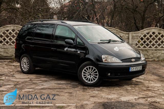 Czarny samochód dostawczy z logo firmy dostarczającej butle gazowe, zaparkowany na zewnątrz, z widocznymi detalami jak numer rejestracyjny i felgi.