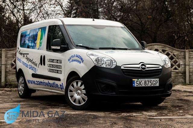 Biało-czarny samochód dostawczy z reklamą butli gazowych i numerami telefonów, zaparkowany na zewnątrz w pochmurny dzień.