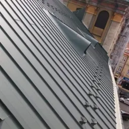 Nowe, metalowe pokrycie dachu kościoła z zamontowanymi uchwytami przeciwśniegowymi i elementami wentylacyjnymi, widok z góry pod kątem.