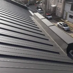 Nowoczesne, grafitowe pokrycie z blachy na dachu budynku mieszkalnego, widok z góry na ułożone panele z widocznymi łączeniami, w tle zabudowa miejska i zaparkowane samochody.