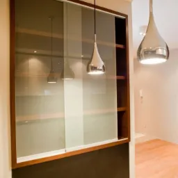 Nowoczesna szafa z przesuwnymi szklanymi drzwiami w drewnianej ramie, oświetlona wiszącymi lampami w kształcie kropli, widok na wnętrze kuchni w tle.