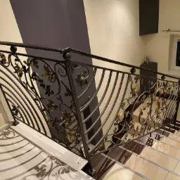 Kute metalowe schody z motywem roślinnym, widok z góry, na stopniach widoczne cienie. Elementy dekoracyjne w kolorze złotym.