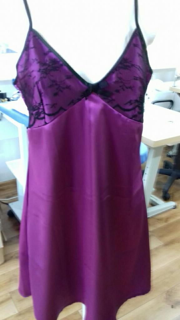 Fioletowa satynowa koszulka nocna z czarną koronką na biuście, sfotografowana w szwalni na tle białego stołu z krzesłem.