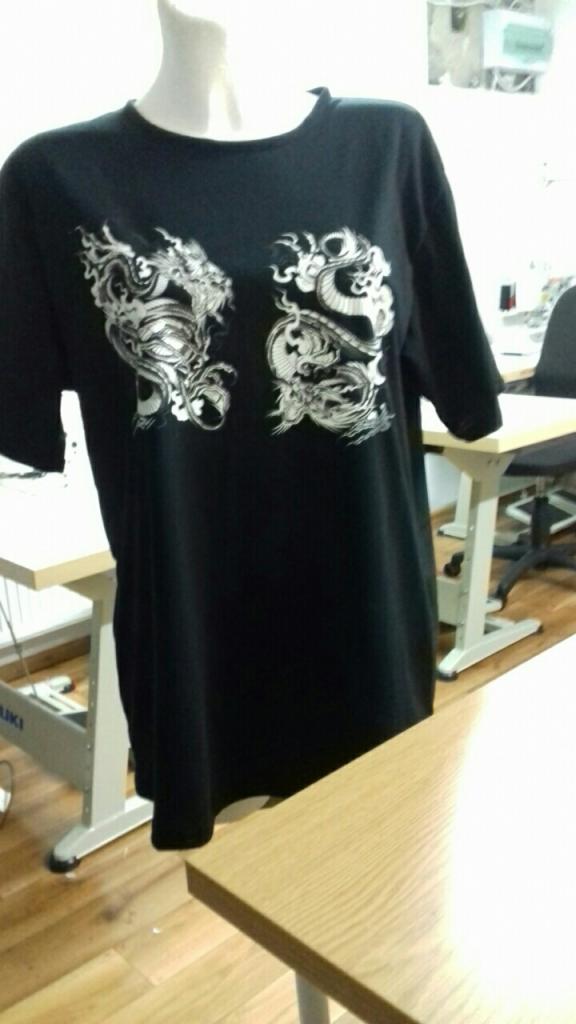 Czarna koszulka z nadrukiem dwóch białych smoków, prezentowana na manekinie w pracowni krawieckiej, z widocznym stołem i maszyną do szycia w tle.