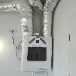 Centrala wentylacyjna z rekuperacją, biała obudowa, podłączona do srebrnych, izolowanych przewodów wentylacyjnych, zamocowana na białej ścianie. Widoczny panel sterowania i etykieta energetyczna.