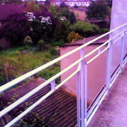 Biała balustrada na balkonie z widokiem na ogród z kwitnącymi krzewami i domy w tle, ujęcie z góry.