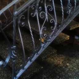 Metalowa balustrada schodowa z ozdobnymi kręconymi elementami, schody wyłożone kamieniami, w tle mur z cegły oraz kartony po prawej stronie.