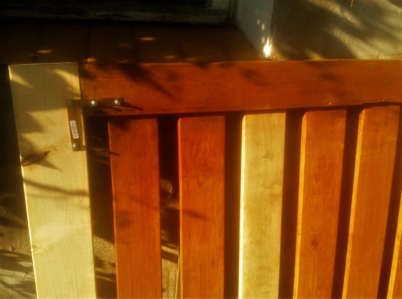 Fragment drewnianej konstrukcji z pionowymi deskami, częściowo pomalowanymi na brązowo, z widocznym metalowym zawiasem, oświetlony ciepłym światłem słonecznym.