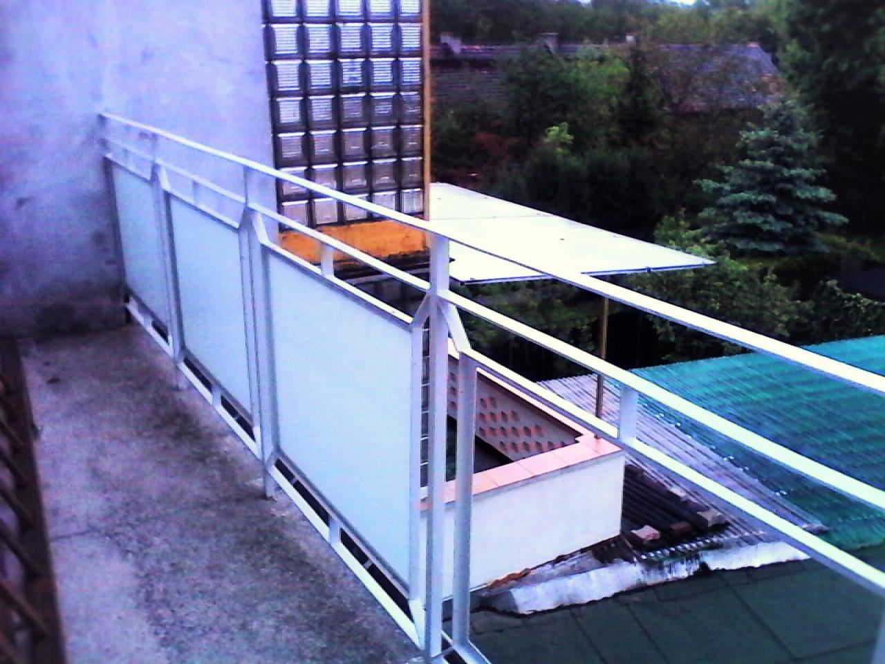 Barierka balkonowa z białymi, matowymi panelami i metalową ramą, widok z góry na otoczenie z zielenią i budynkami w tle.