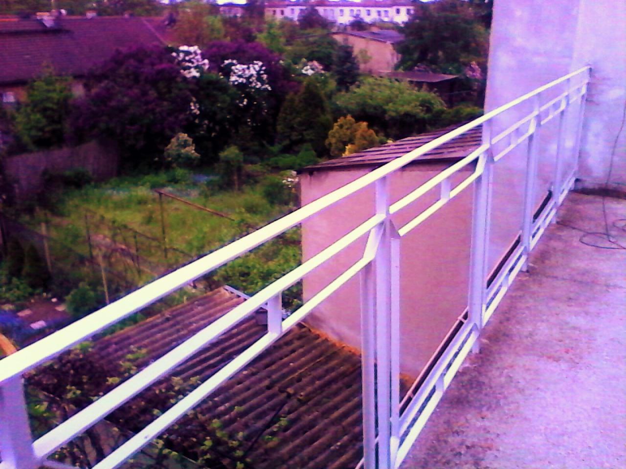 Biała balustrada na balkonie z widokiem na ogród z kwitnącymi krzewami i domy w tle, ujęcie z góry.