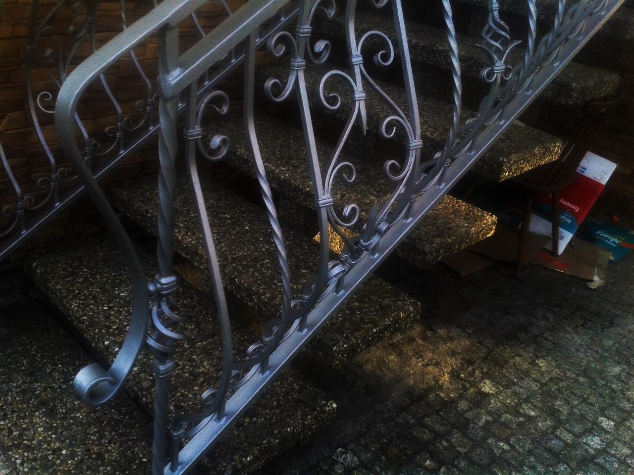 Metalowa balustrada schodowa z ozdobnymi kręconymi elementami, schody wyłożone kamieniami, w tle mur z cegły oraz kartony po prawej stronie.