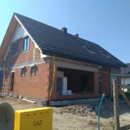 Dom w trakcie budowy z częściowo ukończonym, czarnym dachem z oknami dachowymi, widoczne ceglane ściany i elementy konstrukcyjne, w tle błękitne niebo.