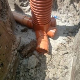 Pomarańczowe rury kanalizacyjne z trójnikiem w wykopie, widoczne połączenia. Instalacja w trakcie montażu, ziemia wokół rur.