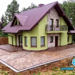 Nowoczesny dom z zieloną elewacją i balkonem, otoczony nowo ułożoną kostką brukową w odcieniach brązu i beżu, z widocznym systemem odprowadzania wody na obrzeżach.