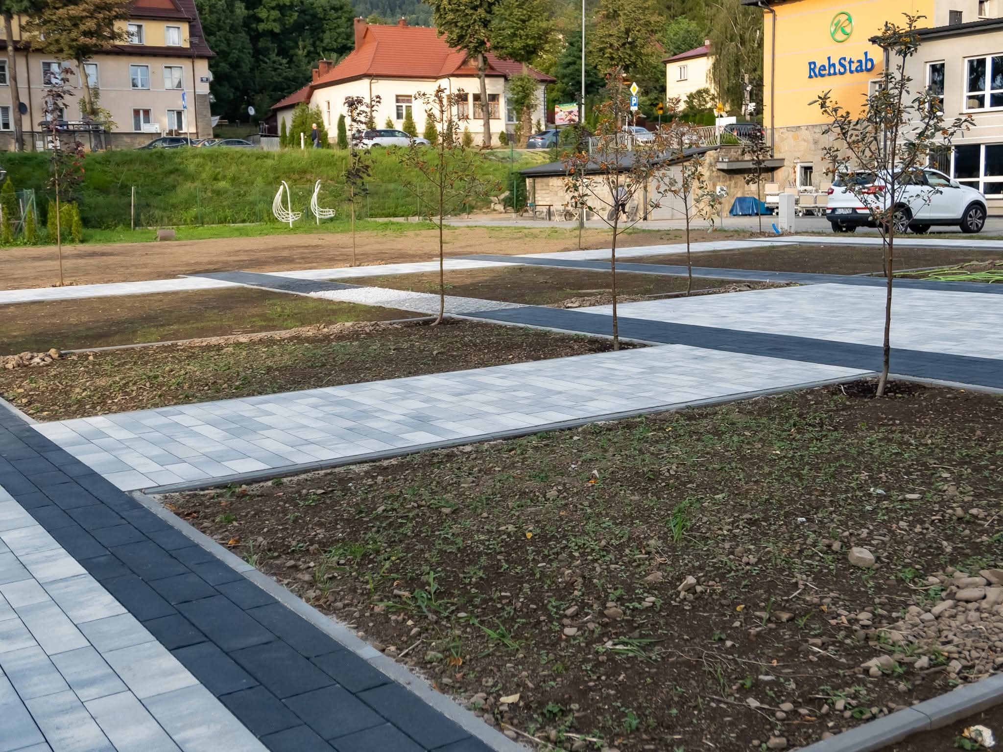 Nowo ułożona, szara kostka brukowa z ciemnymi obrzeżami tworzy ścieżki i obramowania rabat z młodymi drzewkami. Tło stanowi budynek i zieleń.