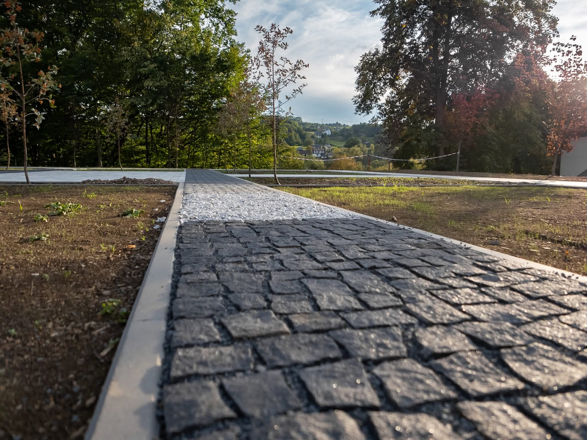 Ścieżka z kostki granitowej w Łukowicy, z białym żwirem po bokach, prowadząca przez park. Widok z niskiej perspektywy, podkreślający fakturę kamienia.