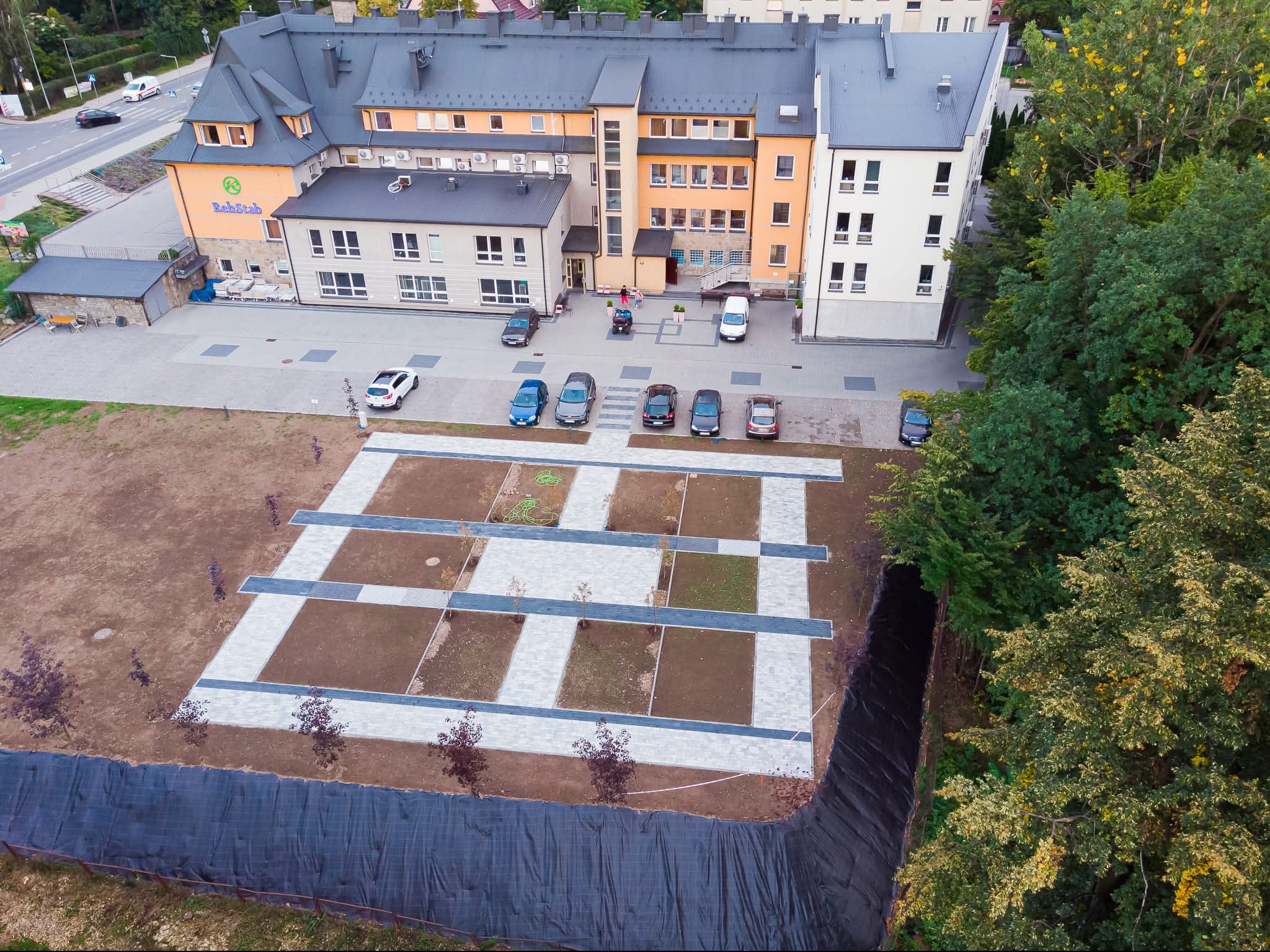 Widok z góry na plac z ułożoną kostką brukową w geometryczny wzór, obok budynku i parkingu. Czarna folia zabezpiecza skarpę. Nowe nasadzenia drzew.