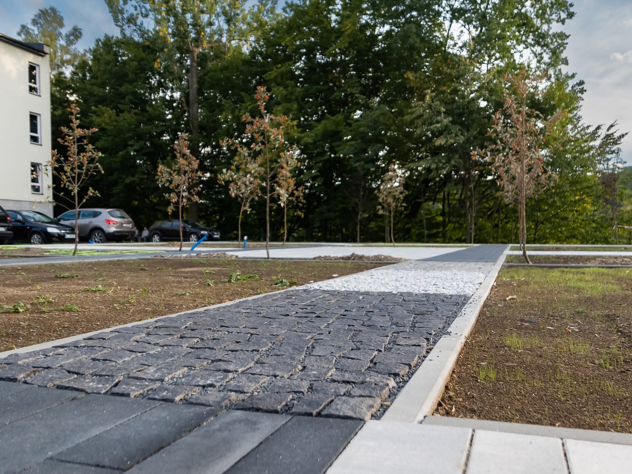 Ścieżka z ciemnej kostki brukowej, obramowana jasnym betonem, prowadząca przez trawnik w kierunku parkingu i budynku. Widoczne drzewa i samochody w tle.
