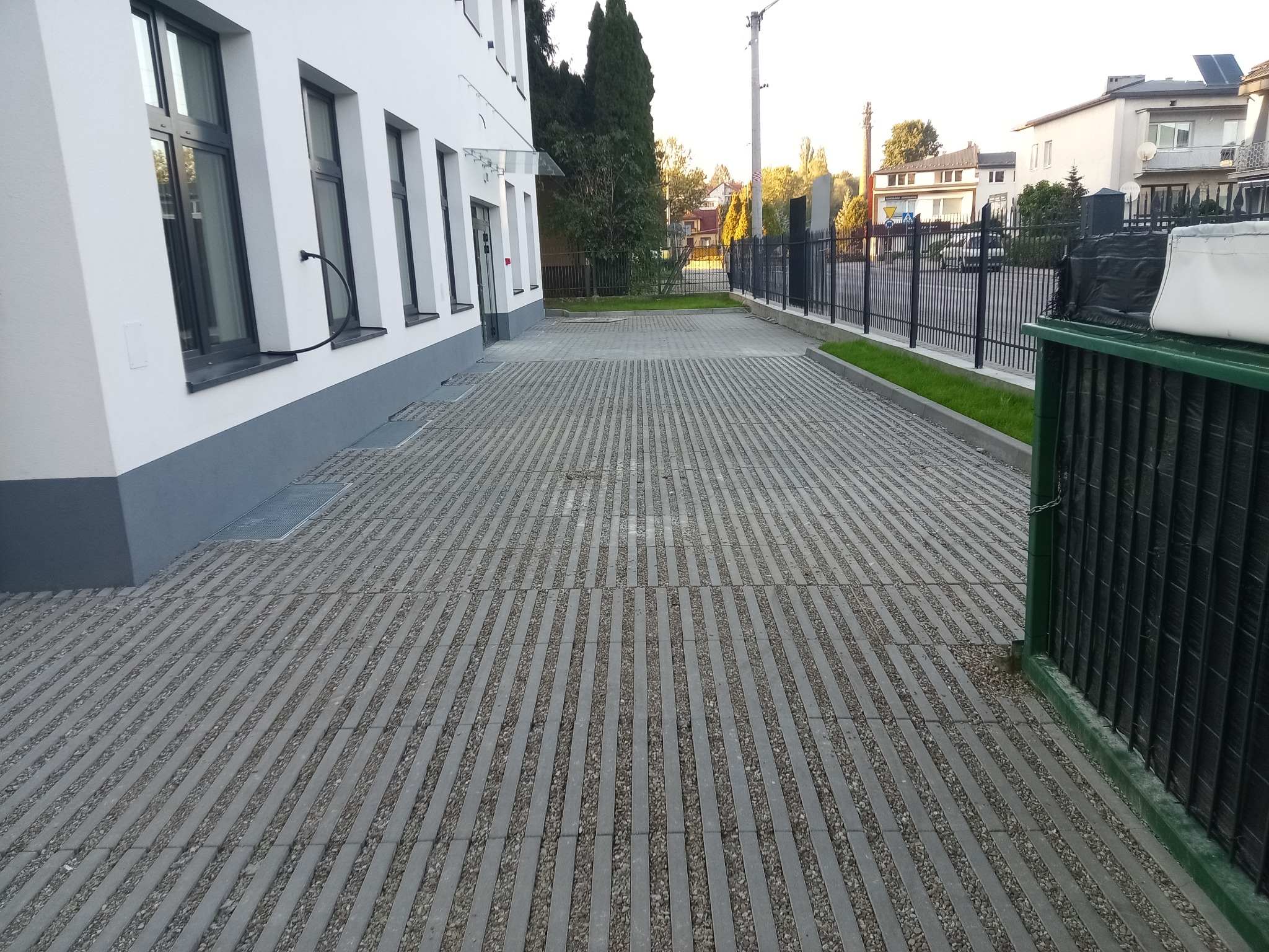 Szara, ażurowa kostka brukowa ułożona wzdłuż budynku z ciemnymi oknami. Widoczny fragment ogrodzenia i zieleń. Ujęcie z perspektywy chodnika.