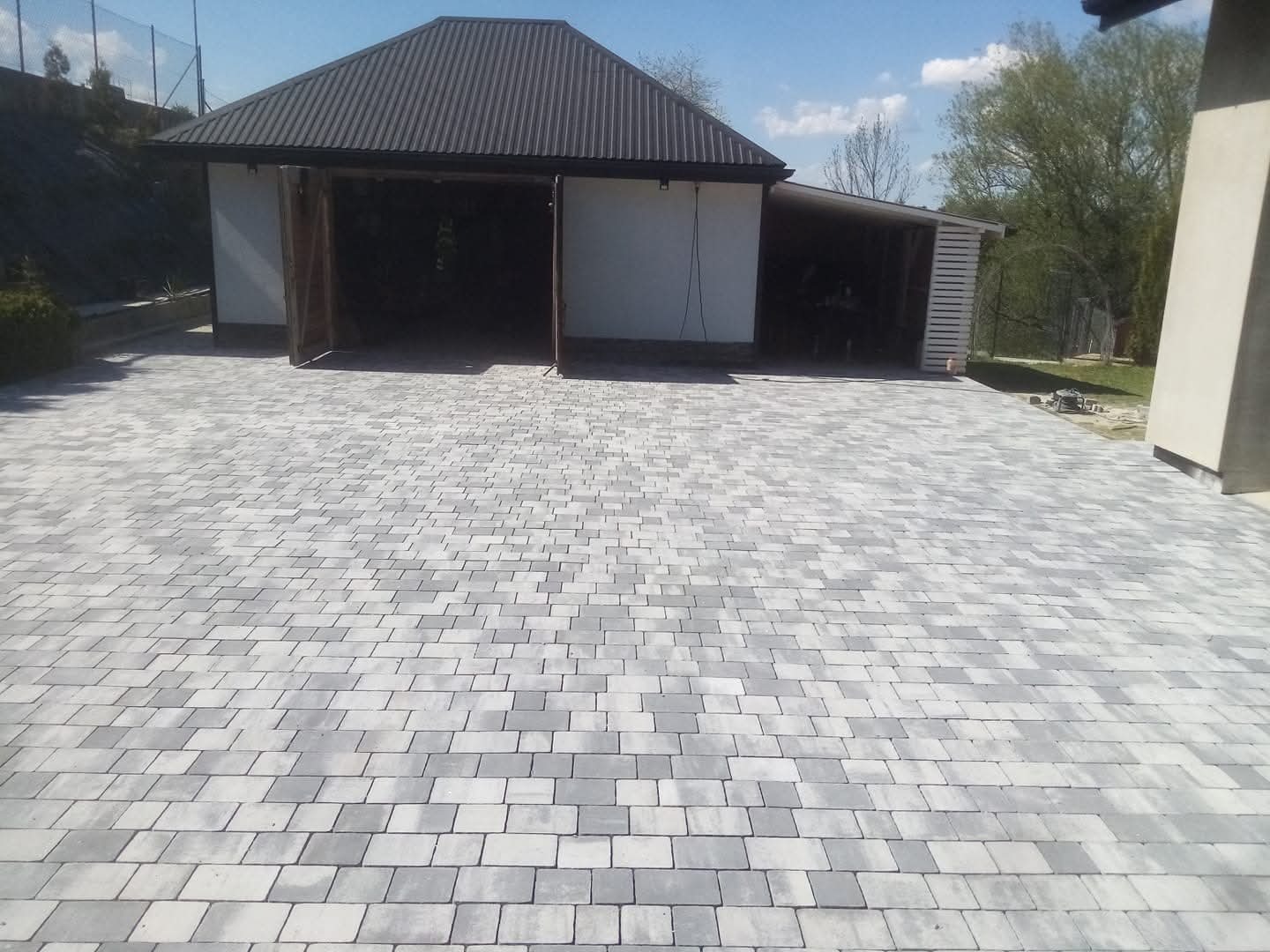 Szara kostka brukowa ułożona na podjeździe przed garażem z otwartymi wrotami. Widok na budynek i fragment zieleni w tle, słoneczny dzień.