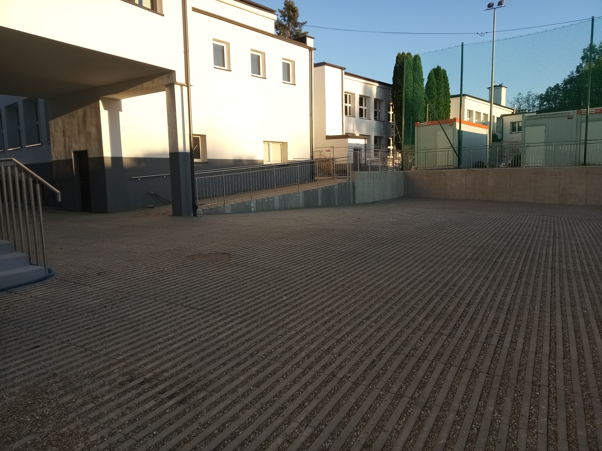 Ułożona, szara nawierzchnia z kostki brukowej na dużym placu przed budynkiem. Widoczne budynki w tle i elementy infrastruktury sportowej.